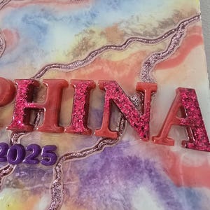 Puede incluir: Una pieza de arte decorativa de resina con el nombre "CHINA" en letras rojas con purpurina. El fondo presenta un dise&ntilde;o abstracto colorido con tonos rosas, azules y morados, y el a&ntilde;o "2025" en morado.