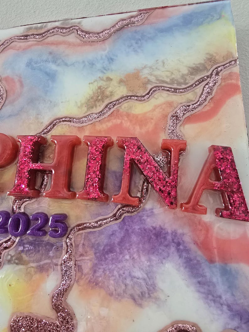 Puede incluir: Una pieza de arte decorativa de resina con el nombre "CHINA" en letras rojas con purpurina. El fondo presenta un dise&ntilde;o abstracto colorido con tonos rosas, azules y morados, y el a&ntilde;o "2025" en morado.
