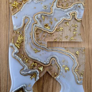 K&ouml;nnte beinhalten: Ein dekorativer Telefonst&auml;nder aus Kunstharz mit einzigartigem Design. Der St&auml;nder hat ein hellblaues und wei&szlig;es Marmormuster mit goldenen und silbernen Glitzerakzenten. Die R&auml;nder sind goldfarben umrandet und der St&auml;nder hat die Form eines stilisierten Buchstabens.