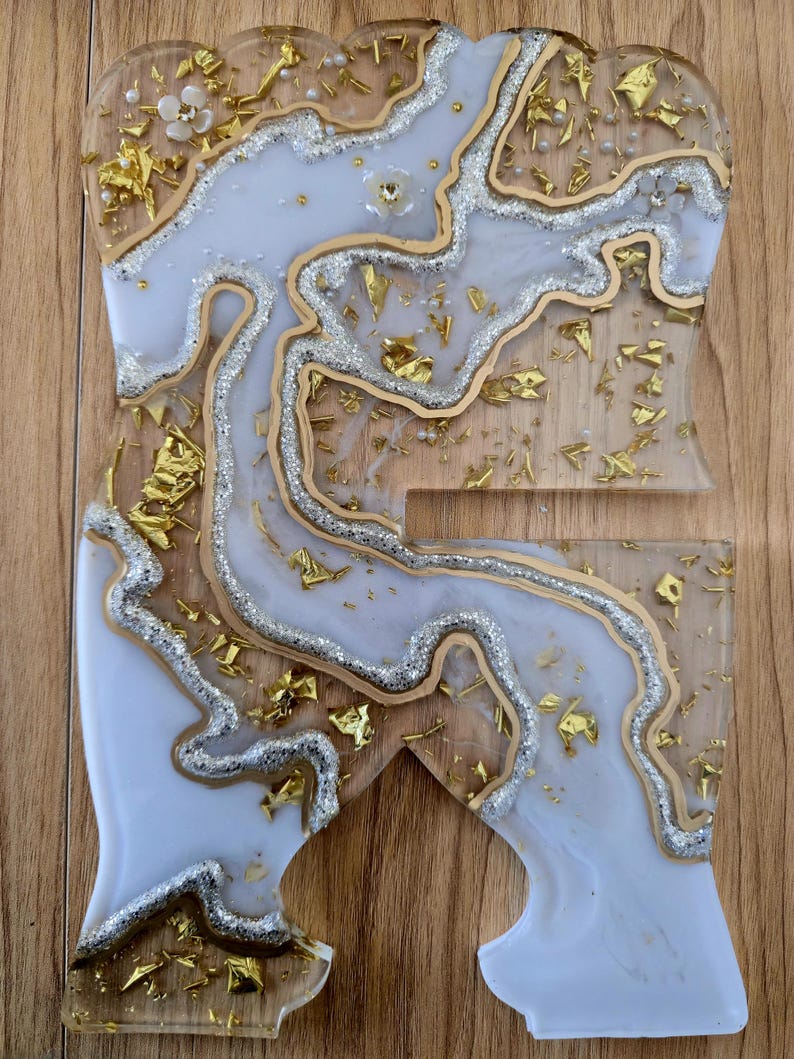 K&ouml;nnte beinhalten: Ein dekorativer Telefonst&auml;nder aus Kunstharz mit einzigartigem Design. Der St&auml;nder hat ein hellblaues und wei&szlig;es Marmormuster mit goldenen und silbernen Glitzerakzenten. Die R&auml;nder sind goldfarben umrandet und der St&auml;nder hat die Form eines stilisierten Buchstabens.