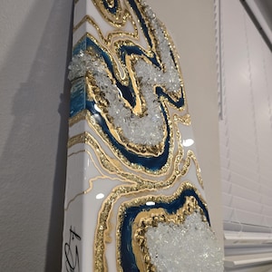 Op de afbeelding: Abstract kunstwerk van hars met een geode-ontwerp. Het kunstwerk heeft witte, gouden en marineblauwe kleuren, met heldere kristalaccenten. Het stuk is aan de zijkant met zwarte inkt gesigneerd.