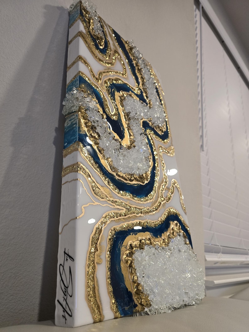 Op de afbeelding: Abstract kunstwerk van hars met een geode-ontwerp. Het kunstwerk heeft witte, gouden en marineblauwe kleuren, met heldere kristalaccenten. Het stuk is aan de zijkant met zwarte inkt gesigneerd.
