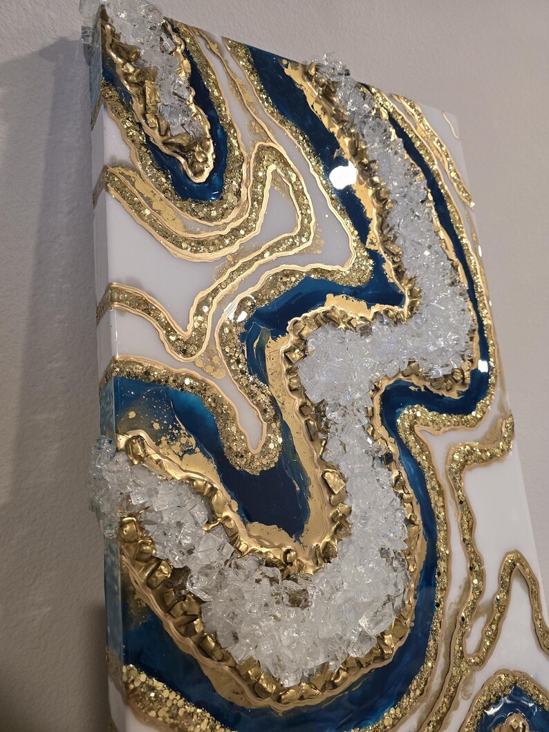 Op de afbeelding: Abstracte wandkunst met een geode-ontwerp. Het kunstwerk bevat witte, marineblauwe en gouden hars, met heldere kristallen accenten. Het stuk heeft een getextureerd oppervlak en een glanzende afwerking, wat een luxe uitstraling cre&euml;ert.