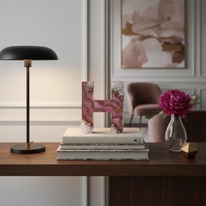 May include: A decorative letter "H" made of pink resin with gold accents and crystal edges. The letter sits atop a stack of books on a wooden surface, next to a black desk lamp and a vase with a pink flower.