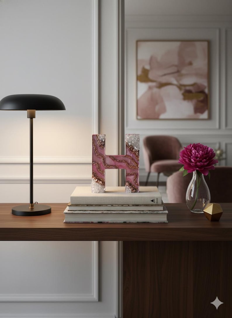 May include: A decorative letter "H" made of pink resin with gold accents and crystal edges. The letter sits atop a stack of books on a wooden surface, next to a black desk lamp and a vase with a pink flower.