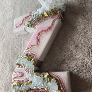 May include: A decorative letter "Z" with a pink and white design, accented with gold glitter and clear crystals. The letter is embellished with a pink glitter outline and topped with a cluster of clear quartz crystals.