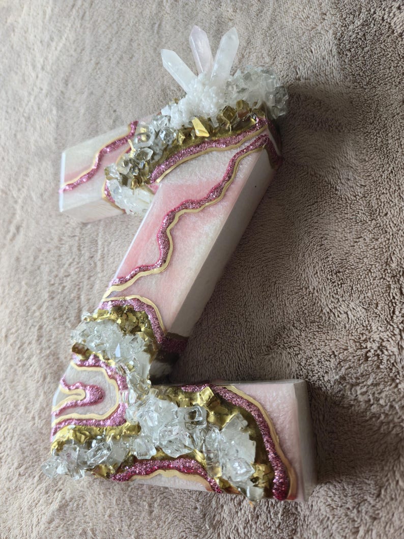 May include: A decorative letter "Z" with a pink and white design, accented with gold glitter and clear crystals. The letter is embellished with a pink glitter outline and topped with a cluster of clear quartz crystals.