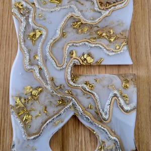 K&ouml;nnte beinhalten: Zweiteiliges Kunstset aus Kunstharz mit wei&szlig;er Basis, silbernen Glitzerakzenten und Goldblatt-Einschl&uuml;ssen. Die Teile haben eine einzigartige Form mit flie&szlig;endem, organischem Design. Auf einer Holzoberfl&auml;che.