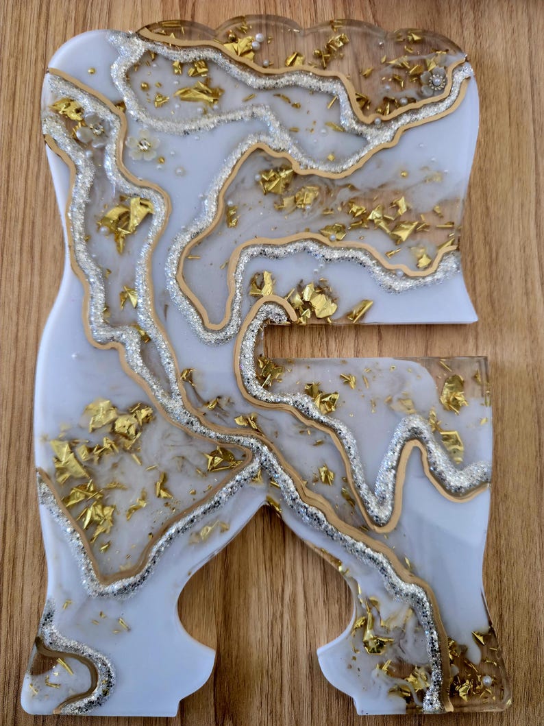 K&ouml;nnte beinhalten: Zweiteiliges Kunstset aus Kunstharz mit wei&szlig;er Basis, silbernen Glitzerakzenten und Goldblatt-Einschl&uuml;ssen. Die Teile haben eine einzigartige Form mit flie&szlig;endem, organischem Design. Auf einer Holzoberfl&auml;che.