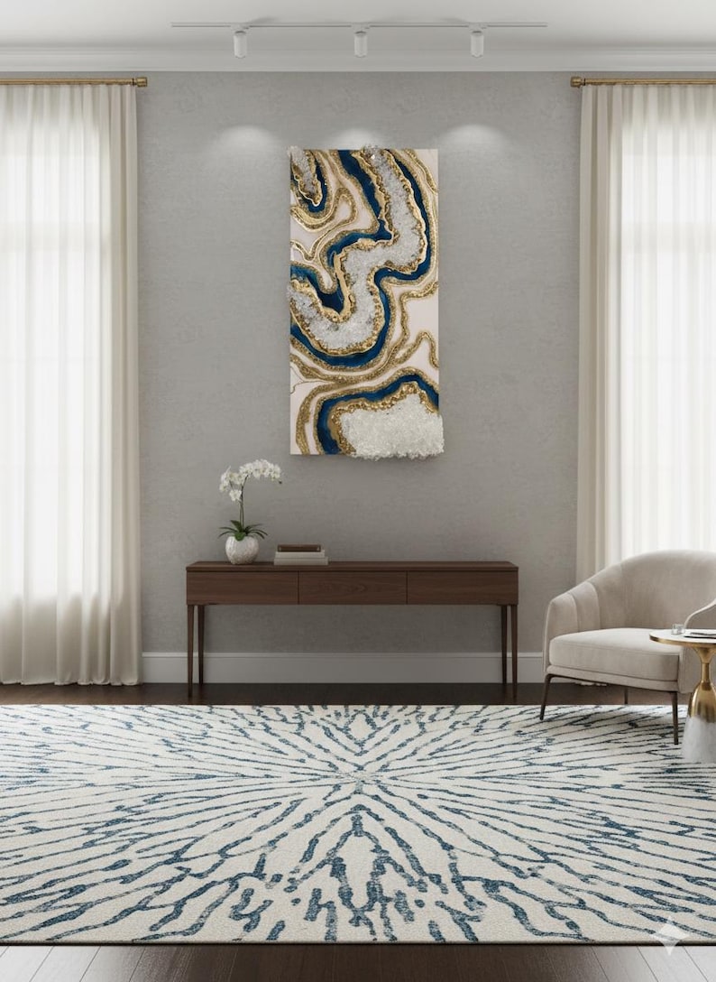 Op de afbeelding: Abstracte wandkunst met een cr&egrave;mekleurige achtergrond, met vloeiende patronen in goud en marineblauw, met een textuurrijk wit kristalachtig element. Het kunstwerk hangt boven een donkerhouten console, met een cr&egrave;mekleurige stoel en een patroonkleed.
