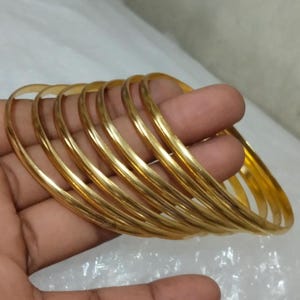 May include: A collection of gold bangles, each with a smooth, polished surface. The bangles are held in a hand, showcasing their circular design and warm, metallic color. The image highlights the jewelry's simple elegance.