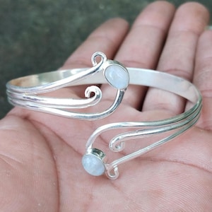 Puede incluir: Un brazalete plateado con dos piedras preciosas redondas de color azul claro. El brazalete presenta un diseño abierto con líneas curvas y remolinos decorativos. Las piedras están engastadas en un engaste de bisel.