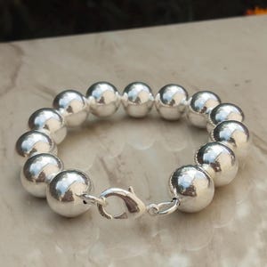 Può includere: Un braccialetto d'argento con grandi perline rotonde e lucide. Le perline sono infilate e fissate con una chiusura d'argento. Il braccialetto è esposto su una superficie chiara.