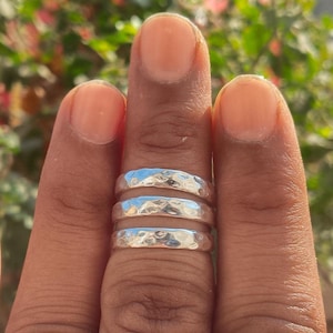 Puede incluir: Tres anillos plateados martillados están apilados en un dedo. Los anillos tienen una superficie texturizada y un aspecto brillante y reflectante. El fondo está borroso, con toques de verde y rosa.