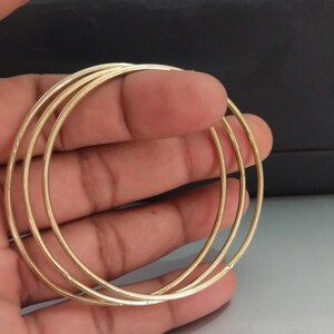 May include: A set of three gold-colored hoop earrings. The earrings are thin and circular, with a smooth, shiny finish. The hoops are held in a hand, with a dark background.
