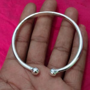 Peut inclure: Un bracelet manchette argenté, ouvert aux extrémités et orné de boules. Le bracelet est présenté sur une main, sur fond rose. Il présente une finition lisse et polie, avec un design simple et élégant, idéal pour un usage quotidien.