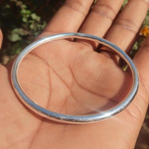 Puede incluir: Un brazalete de color plateado con un diseño liso y redondeado. El brazalete circular parece ser de metal. La imagen muestra el brazalete apoyado en una mano, con un fondo borroso.