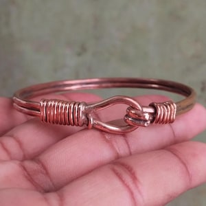 Puede incluir: Una pulsera de color cobre con cierre de gancho. La pulsera presenta un diseño de alambre enrollado a ambos lados del gancho. El gancho es de diseño simple y curvado. La pulsera es de diseño simple y elegante.