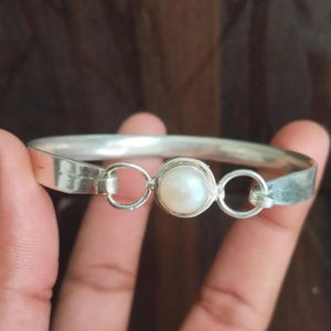 Puede incluir: Una pulsera de plata con una perla blanca redonda en el centro. La pulsera tiene un diseño sencillo con un cierre a cada lado de la perla. La pulsera está sujeta por una persona.
