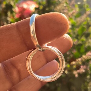 May include: A silver ring with three connected circular bands. The ring is held up by a finger, with a blurred background of green foliage and pink flowers.