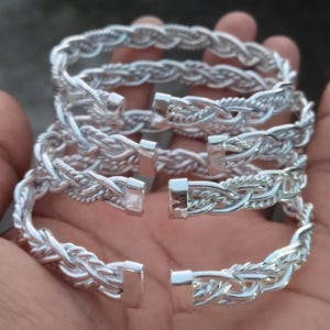 Op de afbeelding: Verschillende zilverkleurige gevlochten armbanden met een sluiting. De armbanden zijn in een stapel gerangschikt, wat hun ingewikkelde geweven ontwerp laat zien. Het metaal heeft een heldere, reflecterende afwerking.