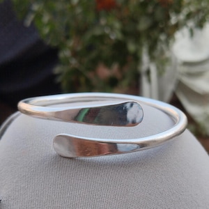 Op de afbeelding: Een zilverkleurige, open manchetarmband. De armband heeft een gladde, gepolijste afwerking met afgeplatte, afgeronde uiteinden. Het ontwerp is een eenvoudige, elegante spiraal, geschikt voor dagelijks gebruik. De armband heeft een diameter van ongeveer 6,35 cm.