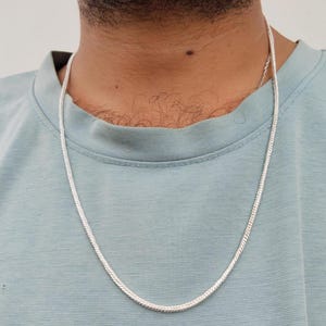 May include: A silver-toned chain necklace with a smooth, rounded, and tightly linked snake-style design. The chain rests against a light blue t-shirt, featuring a polished metallic finish that reflects light along its continuous, flexible length.