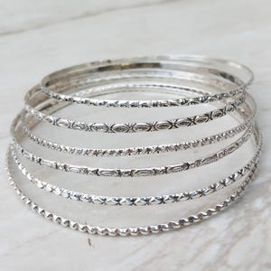 May include: A stack of silver bangles with various etched patterns. The bangles are thin and circular, with designs including floral, geometric, and textured lines. The jewelry is displayed on a light-colored, marble-like surface.