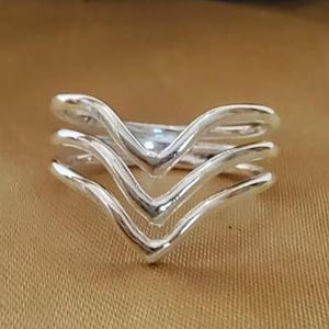 May include: A silver ring with a layered chevron design. The ring features three stacked V-shaped bands, creating a modern and geometric look. The ring is displayed on a gold-colored surface.