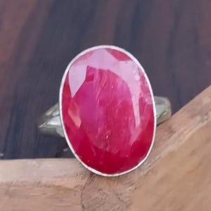 May include: A silver ring with a large, oval-cut ruby gemstone. The ruby is a deep red hue and is set in a plain silver band. The ring is shown on a wooden surface.