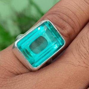 May include: A rectangular, aquamarine-colored gemstone ring with a silver band. The gemstone is a vibrant teal color and is set in a simple, polished silver setting. The ring is shown on a finger.