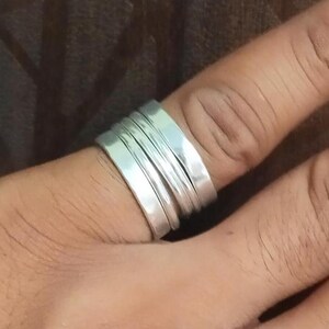 Puede incluir: Un anillo de plata con un diseño apilado se lleva en un dedo. El anillo tiene múltiples bandas, creando un efecto en capas. El metal tiene un acabado pulido, que refleja la luz. El anillo es una joya.