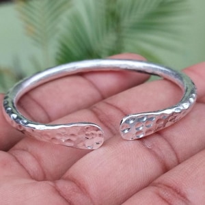 Könnte beinhalten: Ein silberfarbenes, offenes Armreif-Armband mit gehammerter Textur. Das Armband wird auf einer Hand gezeigt, wobei die offenen Enden zum Betrachter zeigen. Die gehammerte Textur verleiht dem Armband ein einzigartiges, handgefertigtes Aussehen.