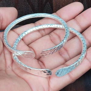 May include: Two silver-colored, open-ended bracelets with a hammered texture. The bracelets are resting on a hand, showcasing their simple, yet elegant design. The open ends allow for easy adjustment and wear.