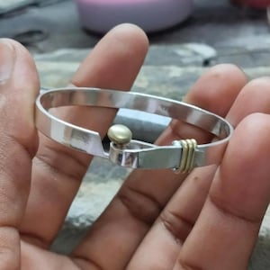 Könnte beinhalten: Ein silberfarbenes Armband mit Hakenverschluss. Das Armband hat eine kleine goldfarbene Perle und goldfarbene Drahtakzente. Das Armband wird in der Hand einer Person gehalten.