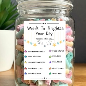 May include: A glass jar filled with colorful rolled-up paper notes. A label reads "Words To Brighten Your Day" with prompts like "Need Confidence" and "Feel Anxious." The jar is on a wooden surface.
