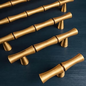 May include: Gold-colored bamboo-style cabinet pulls arranged on a dark blue surface. The pulls have a textured, segmented design, resembling bamboo stalks. Each pull is mounted on a small, triangular base, providing a decorative and functional hardware option.