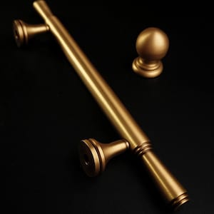 May include: A set of gold-coloured door hardware, including a long handle with two mounting points and a round knob. The hardware is made of metal and has a matte finish. The items are displayed on a black background.
