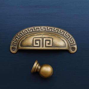 May include: Antique brass-toned drawer pulls and knob. The half-moon pull features a Greek key design and a central square pattern. The knob is a simple, round design. Ideal for furniture restoration or new builds.