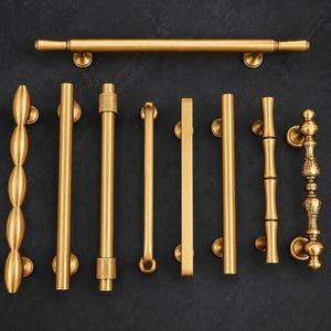 Può includere: Una collezione di maniglie per porte e armadi color ottone in vari design. Gli stili includono una barra lunga, un rametto, una serie di forme ovali e diverse maniglie a barra verticale. Le maniglie sono esposte su uno sfondo scuro.