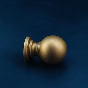 May include: A gold-colored, spherical curtain rod finial with a cylindrical base. The finial is made of metal and has a matte finish. The background is a dark blue.