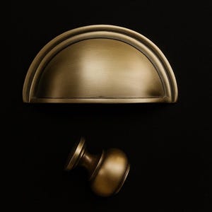 Puede incluir: Dos herrajes de latón antiguo para armarios sobre un fondo negro. Uno es un tirador de media luna y el otro un pomo redondo. El herraje tiene un acabado cálido y envejecido, lo que sugiere un estilo vintage o clásico. Ideal para armarios de cocina o baño.