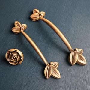 May include: Two bronze-colored cabinet handles and a matching knob. The handles have a curved design with leaf-shaped accents at each end. The knob features a rose design. All pieces are made of metal.