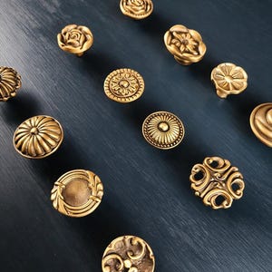 May include: A collection of antique brass cabinet knobs with floral and geometric patterns. The knobs are displayed on a dark blue surface, highlighting their detailed designs and warm, metallic finish. These decorative knobs are ideal for adding a vintage touch to furniture.