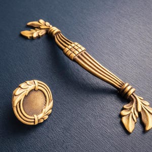 May include: A set of antique brass cabinet hardware, including a long handle and a round knob. The handle features a ribbed design with leaf accents, while the knob has a laurel wreath border. The hardware is set against a dark blue background.