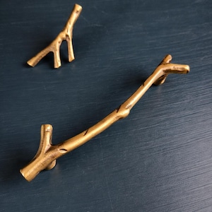May include: Two brass-colored twig-shaped cabinet pulls and a small decorative twig on a dark blue surface. The larger pull is horizontal, with a branch-like design. The smaller twig is vertical, with a similar design.