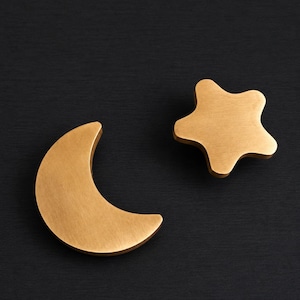 Puede incluir: Dos tiradores decorativos de color latón. Uno tiene forma de media luna y el otro de estrella de cinco puntas. Ambos están colocados sobre un fondo oscuro. Estos tiradores podrían usarse para cajones o armarios.