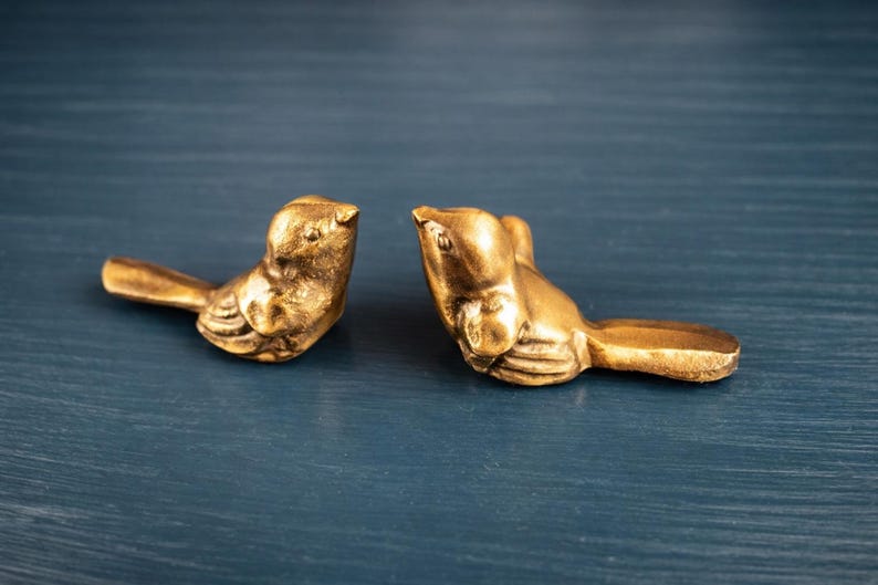 May include: Two gold-colored bird figurines. The birds are facing each other, with detailed wings and tail feathers. The figurines are on a dark blue surface, creating a contrast with the metallic finish.