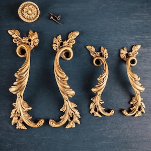 May include: A set of four gold-toned metal cabinet pulls with ornate, swirling leaf-like scrollwork designs, accompanied by a single circular knob featuring a central floral motif and two small metal mounting screws for installation.