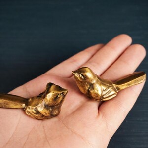 May include: Two small, gold-colored bird figurines. The birds are detailed with visible wings and beaks. They are resting on a person's open palm, with a dark blue background. The birds appear to be made of metal.